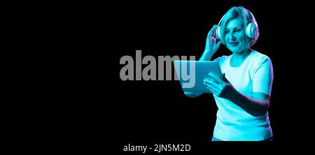 Femme émotif senior en t-shirt blanc utilisant une tablette numérique isolée sur fond sombre dans la lumière du néon. Concept de beauté, âge, émotions, moderne Banque D'Images