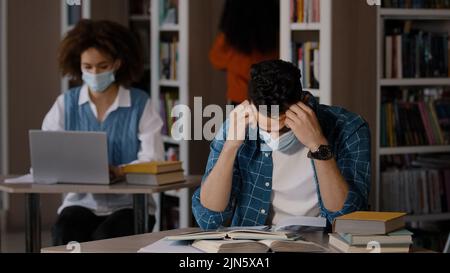 Un jeune homme arabe fatigué surtravaillé dans un masque de protection assis à un bureau dans la bibliothèque fait des devoirs écrit des notes prépare pour l'examen fait des erreurs saisit Banque D'Images