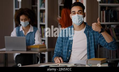 Étudiants dans la bibliothèque jeune indien gars dans le masque de protection assis au bureau faisant des devoirs préparation à l'examen écrire des notes heureux homme regardant l'appareil photo Banque D'Images