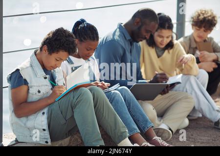 Divers groupes d'enfants assis en rangée et écrivant dans des carnets à l'extérieur pendant la leçon d'été Banque D'Images