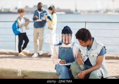 Portrait de deux adolescents noirs écrivant dans des carnets pendant les cours en plein air à la lumière du soleil avec un enseignant en arrière-plan, espace de copie Banque D'Images