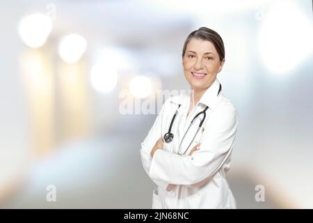 Femme mature médecin dans un manteau blanc de laboratoire, souriant amical, sur le fond flou d'un centre médical intérieur. Banque D'Images