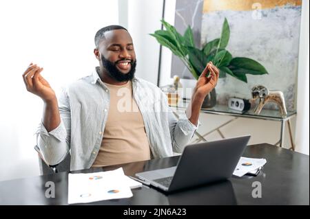 Satisfait calme paisible Afro-américain élégant homme, indépendant, concepteur, pdg de la société, prendre une pause, repos de travail en ligne ou d'étude dans un ordinateur portable, méditant avec les yeux fermés, relaxant, souriant Banque D'Images