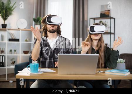 Un jeune couple assis à la table, utilisant des micro-casques VR avec un ordinateur portable moderne pour une expérience de la réalité virtuelle. Une famille heureuse passe du temps libre à la maison et à jouer à des jeux. Banque D'Images