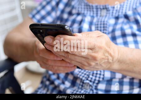 Téléphone mobile dans les mains froissées de femme de près, femme âgée dans la robe damée assis avec un smartphone. Concept de communication en ligne à la retraite Banque D'Images