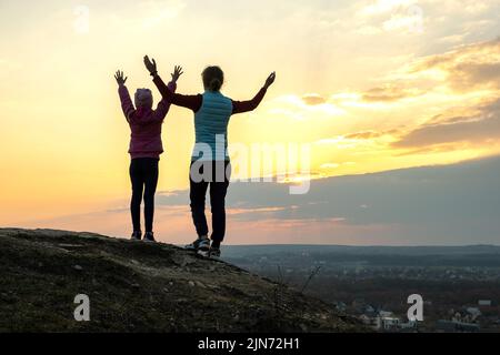 Vue arrière de la jeune femme et de l'enfant fille debout sur une colline herbeuse de montagne avec les bras levés au coucher du soleil appréciant la vue sur la nature. Traditions de tourisme familial Banque D'Images