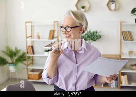 Femme d'affaires senior parlant sur un téléphone à haut-parleur, enregistrant un message vocal ou utilisant un assistant virtuel Banque D'Images