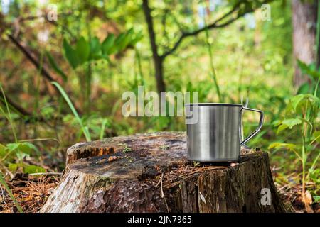 Mug touristique en métal avec boisson chaude se trouve sur une souche dans la forêt. Banque D'Images