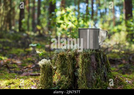 Mug touristique en métal avec boisson chaude se trouve sur une souche dans la forêt. Banque D'Images