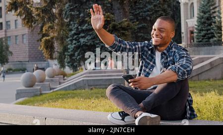 Kharkov, Ukraine - 08.09.2021: Joyeux homme afro-américain assis en ville à l'extérieur regardant le téléphone mobile regarde à distance en signe de salut bonjour Banque D'Images