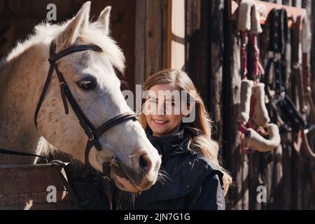 Jeune femme debout à côté du cheval arabe blanc reposant dans une boîte d'écuries en bois, le soleil brille sur eux, détail de gros plan Banque D'Images