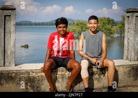 Deux adolescents s'assoient sur un mur de mer souriant, un garçon avec une cigarette, sur fond de mer et d'île tropicale thaïlandaise Banque D'Images