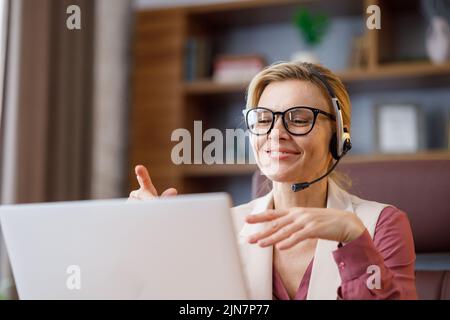 Femme d'affaires souriante portant un casque parlant à la webcam d'un ordinateur portable, au bureau. Enregistrement de vlog, visioconférence en ligne, appel vidéo, en ligne Banque D'Images