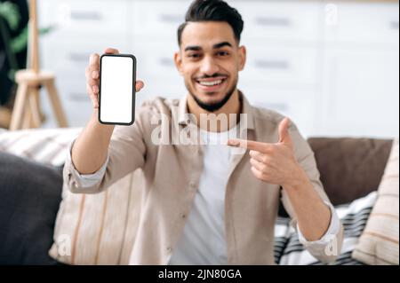 Maquette d'écran du téléphone. Un homme de course mixte souriant et dénature, montrant un smartphone avec un écran maquette blanc vide, un espace pour la publicité ou la présentation, assis sur un canapé dans un salon Banque D'Images