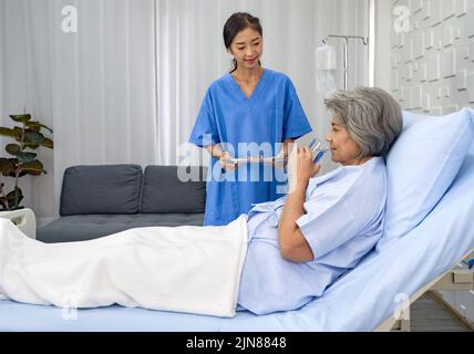 Un patient asiatique âgé à poil blanc reçoit un médicament et un verre d'eau de la part d'une infirmière. Une femme âgée étant traitée à l'hôpital, allongée sur le lit Banque D'Images