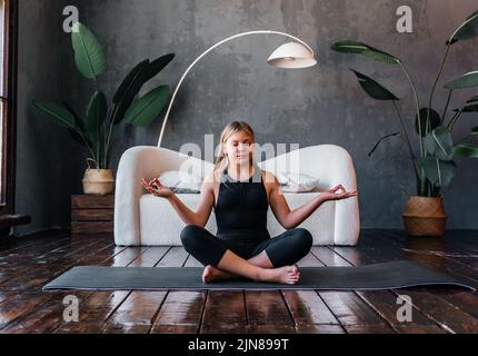 Jeune fille attrayante pratiquant le yoga, assis dans la moitié de l'exercice de Lotus, Ardha Padmasana pose, exercice, session de méditation, intérieur de maison. Quali élevé Banque D'Images