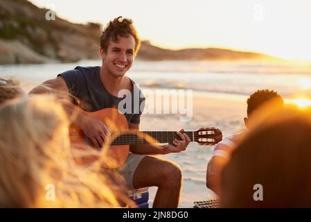 La musique et l'océan sont vraiment tout ce dont nous avons besoin. Un homme jouant de la guitare tout en étant assis sur la plage avec ses amis. Banque D'Images