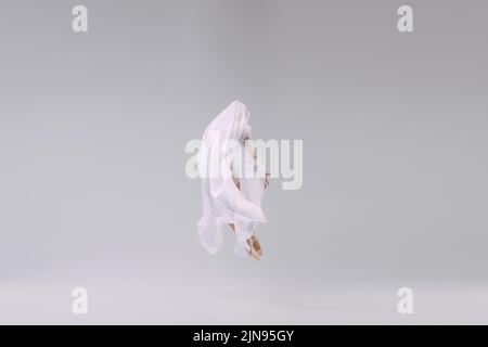 Portrait de la jeune ballerine dansant, sautant entièrement couvert de tissu transparent isolé sur fond gris studio Banque D'Images