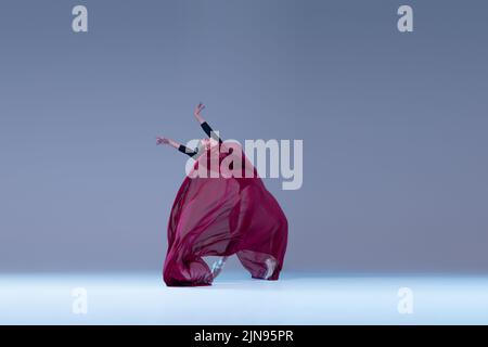 Portrait de jeune danse de ballerine gracieuse avec un tissu rouge profond isolé sur fond bleu gris studio. Performance charmante Banque D'Images