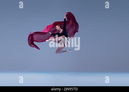 Portrait de la jeune ballerine dansant avec un tissu rouge profond isolé sur fond bleu gris studio. Des performances exceptionnelles Banque D'Images