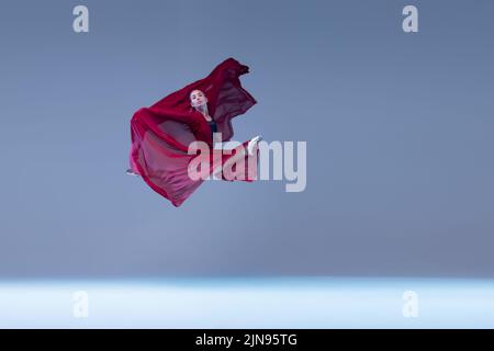 Portrait de la jeune ballerine dansant recouvert d'un tissu rouge foncé transparent isolé sur fond bleu gris studio. Voler haut Banque D'Images