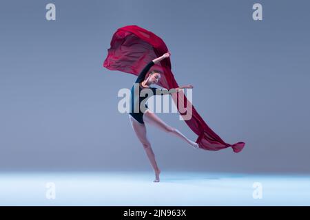 Portrait de la jeune ballerine dansant avec un tissu rouge profond isolé sur fond bleu gris studio. Onde tendre Banque D'Images