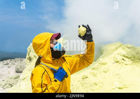 une femme volcanologue, à l'arrière-plan d'un fumarale fumeur, examine un échantillon de minerai de soufre Banque D'Images