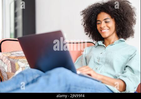 Jeune femme africaine étudiante ou indépendante en tenue décontractée assise sur le canapé et en utilisant un ordinateur portable, travaillant sur le projet, regardant l'écran avec le sourire de plaisir, la websurf ou le shopping en ligne Banque D'Images