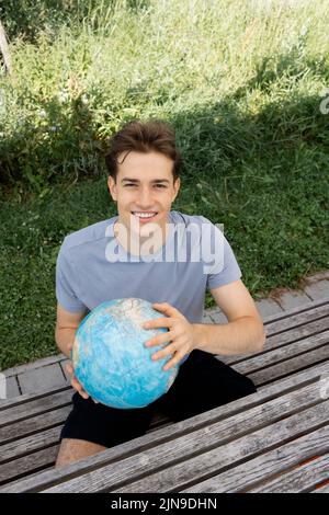 adolescent avec chemise grise assis sur un banc de parc tenant le globe dans sa main et rêvant d'un voyage, des vacances Banque D'Images