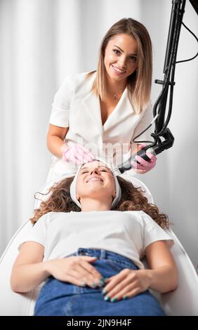 Portrait d'une femme cosmetologiste souriante effectuant le resurfaçage de la peau au laser. Client heureux en prévision du rajeunissement de la peau après une procédure importante. Banque D'Images