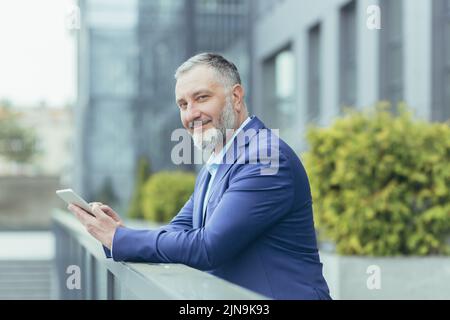 Portrait d'un patron d'homme d'affaires à cheveux gris, homme à l'extérieur du bureau utilisant un ordinateur tablette souriant et regardant l'appareil photo, investisseur en costume d'affaires, banquier heureux Banque D'Images