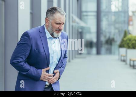 Homme d'affaires senior aux cheveux gris à l'extérieur du bâtiment de bureau, homme en costume d'affaires malade a du mal d'estomac. Banque D'Images