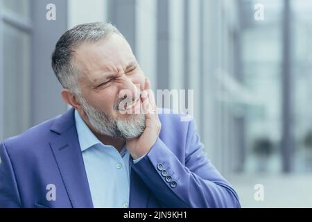 Portrait en gros plan d'un homme d'affaires à cheveux gris, d'un homme d'affaires expérimenté à l'extérieur du bâtiment de bureau, ayant de graves maux de dents Banque D'Images