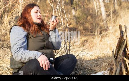 Photo d'une femme assise avec des jambes croisées à l'extérieur le jour d'hiver ensoleillé près du feu de joie regardant la tasse de thé chaud dans sa main. Banque D'Images
