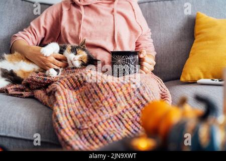 Femme en écossais buvant le thé chaud de Halloween thème mug, en train de caresser l'animal de compagnie de chat multicolore décontracté sur le canapé à la décoration pour l'automne maison de vacances. Confortable Banque D'Images
