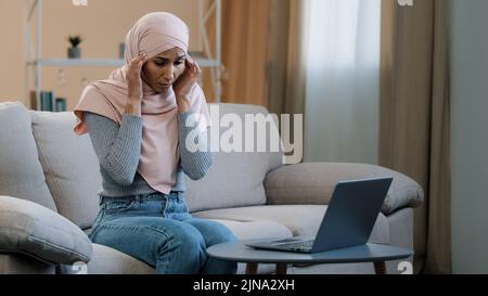 Jeune femme musulmane en hijab rose assis sur le canapé en utilisant un ordinateur portable ayant mal de tête migraine triste femme freelancer se sentent la douleur sévère fatiguée stressée Banque D'Images