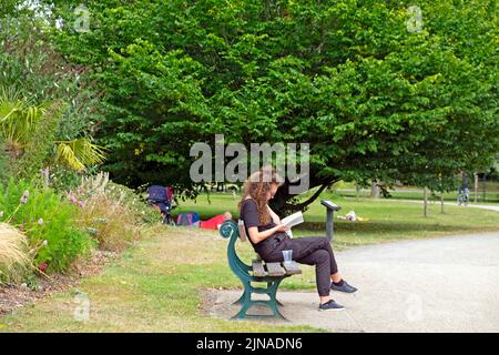 Jeune femme assise sur le banc du parc lisant un livre par des arbres en été août 2022 Bute Park Cardiff pays de Galles Royaume-Uni Grande-Bretagne KATHY DEWITT Banque D'Images