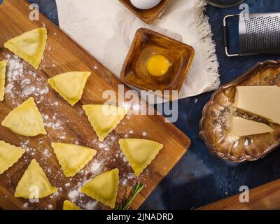 Raviolis italiens faits maison sur une planche à découper en attente de cuisson. Ingrédients, ustensiles de cuisine sur fond bleu foncé. Recettes pour la cuisson de raviolis Banque D'Images