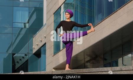 Forte femme calme musulmane fille islamique dans hijab pieds nus dame dans le pantalon violet se tient ville bâtiment arrière-plan fléchissant le genou tire la hanche arrière et devant Banque D'Images