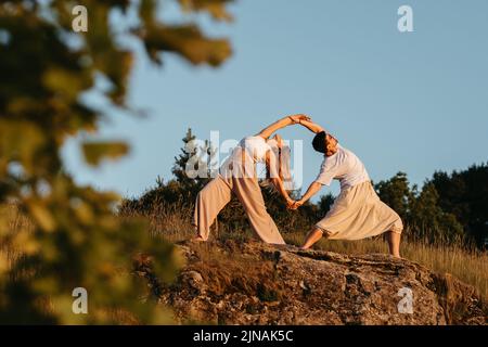 Jeune couple adulte faisant une belle pose, homme et femme pratiquant le yoga à l'extérieur sur la Rocky Hill au coucher du soleil Banque D'Images