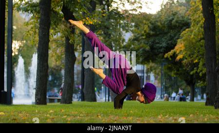 Forte active musulmane fille dans hijab femme yogi sportswoman faire l'entraînement de yoga dans le parc sur pelouse verte faire l'équilibre entraînement de main tenant la jambe dans l'air Banque D'Images