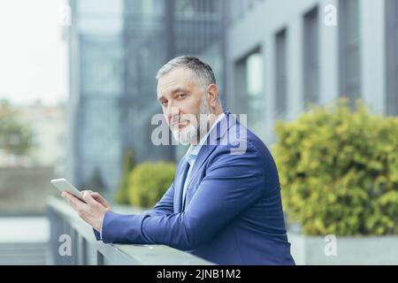 Portrait d'un investisseur sérieux et concentré, senior et expérimenté, un homme aux cheveux gris à l'extérieur du bureau regardant attentivement l'appareil photo, homme d'affaires en costume tenant une tablette informatique Banque D'Images