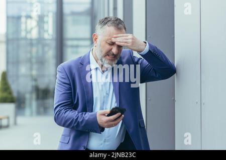 Un homme d'affaires aux cheveux gris est malade à l'extérieur de l'immeuble de bureaux, un homme tenant la main à la tête, ayant des maux de tête graves, un patron essayant d'appeler un médecin au téléphone et d'appeler une ambulance Banque D'Images