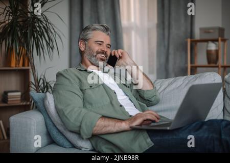 Joyeux homme européen mûr avec des appels à la barbe par téléphone, travaille sur ordinateur portable, est assis sur un canapé dans le salon intérieur Banque D'Images