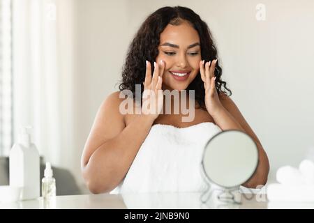 Concept de soin du visage et de la peau. Jeune femme noire de taille plus massant son visage et regardant le miroir Banque D'Images
