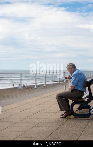Un vieil homme assis sur un banc près de la mer tenant un bâton de marche avec ses yeux fermés Banque D'Images