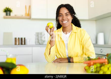 Régime de perte de poids. Bonne dame noire tenant la pomme et souriant à l'appareil photo, assis dans l'intérieur moderne de la cuisine, espace de copie Banque D'Images