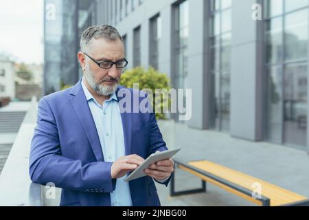 Banquier principal concentré et sérieux à cheveux gris utilisant une tablette informatique à l'extérieur du bâtiment de bureau, homme d'affaires lisant les nouvelles et la pensée, homme dans des lunettes et costume d'affaires tapant le message Banque D'Images