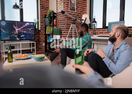 Jeune adulte utilisant le joystick et la console pour jouer à des jeux vidéo à la maison se réunissant avec des amis, boire de la bière et manger des collations. Groupe de personnes s'amusant avec la compétition de jeu, le divertissement. Banque D'Images