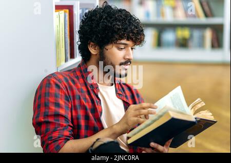 Lecture de livres. Étudiant indien ou arabe spécialisé dans l'université ou l'université, assis dans une bibliothèque près d'une étagère, lisant un livre intéressant, se préparant à l'examen, faisant des devoirs, acquérir des connaissances Banque D'Images
